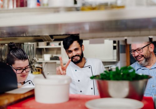 Drei Männer stehen in der Küche, der mittlere zeigt ein Peace-Zeichen